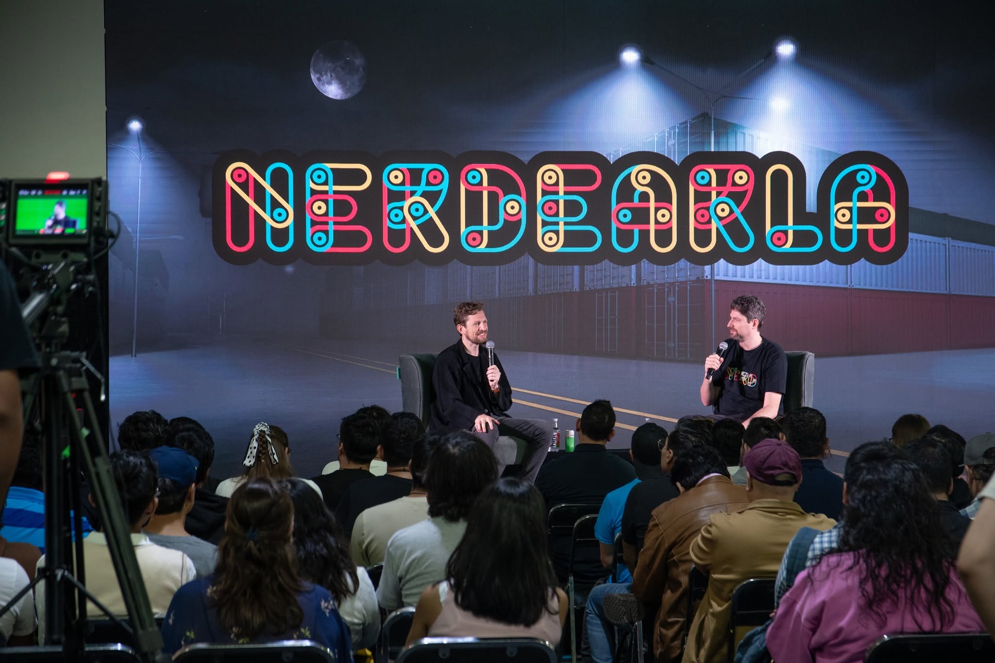 A two-person conversation under the glowing Nerdearla sign, in front of a full room